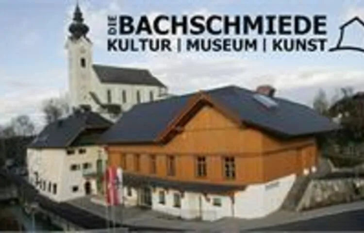 Vista aérea del edificio del centro cultural Die Bachschmiede con fachada naranja y una torre de iglesia que se alza detrás, con el texto superpuesto del nombre del lugar que dice 'DIE BACHSCHMIEDE KULTUR | MUSEUM | KUNST'