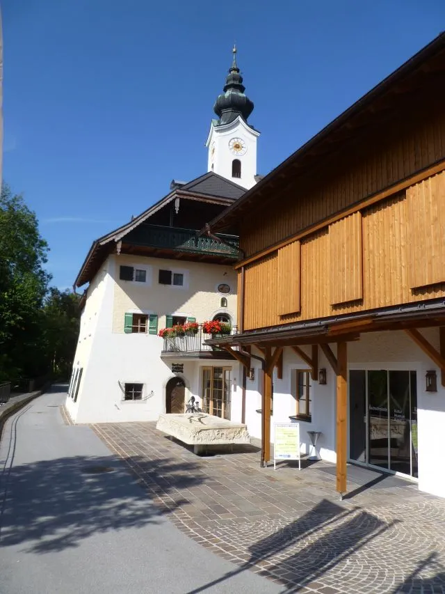 Vista exterior del centro cultural Die Bachschmiede que muestra un edificio alpino blanco con un balcón adornado con macetas de flores, una estructura anexa de madera y una torre de iglesia con cúpula de cebolla verde bajo un cielo azul despejado.