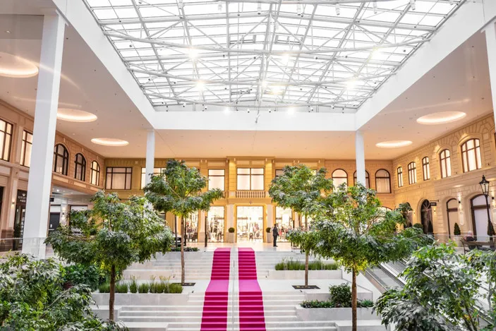 Przestronne atrium z przeszklonym sufitem, białymi kolumnami i różowym dywanem prowadzącym do wejścia do budynku otoczonego zielonymi drzewami i roślinami.
