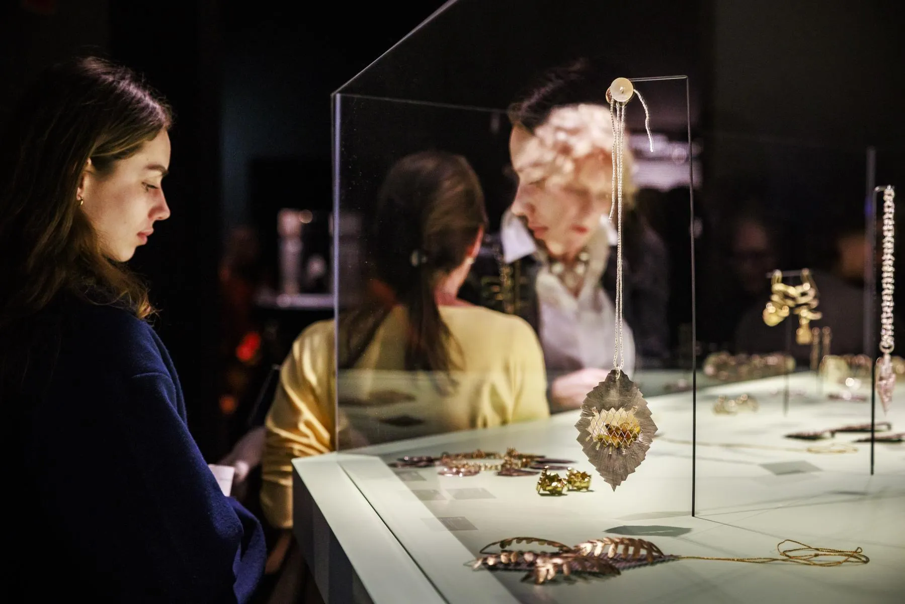 Museum visitors viewing jewelry and pendants displayed inside a glass case with reflections visible on the glass.