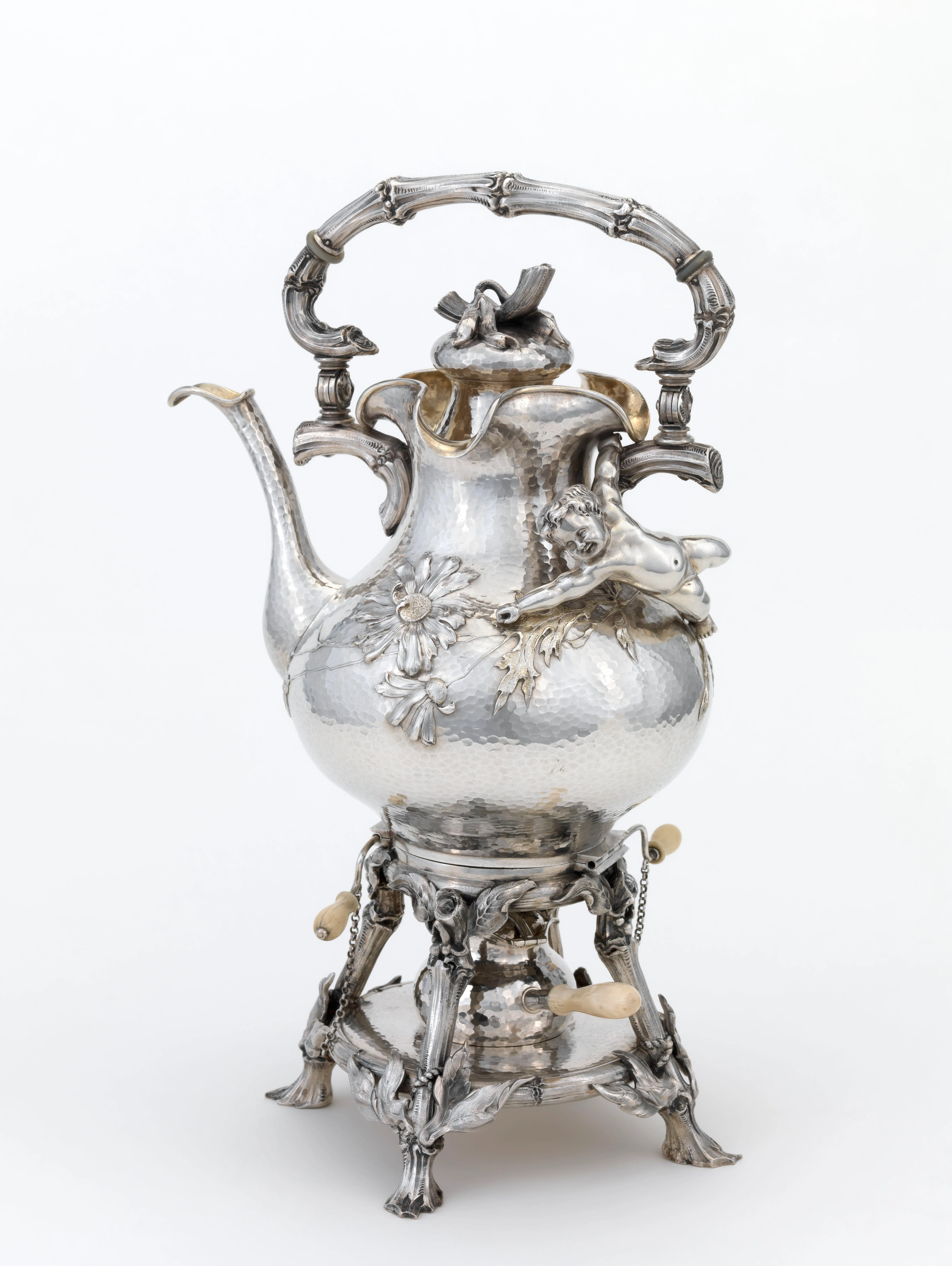 Tetera de plata ornamentada sobre un hornillo, con asa de estilo bambú, una figura de rana, decoraciones en relieve floral y pomos con punta de marfil en la base