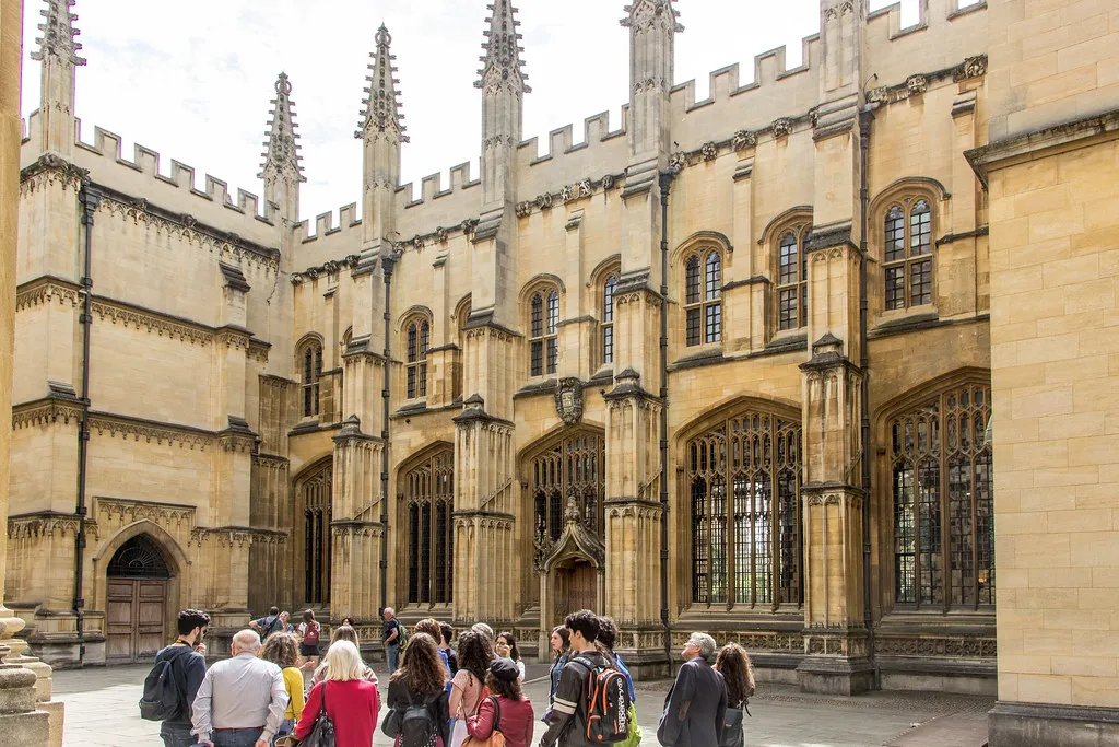 Gothic stone facade of the Divinity School with ornate pinnacles, arched windows, and visitors gathered in the courtyard