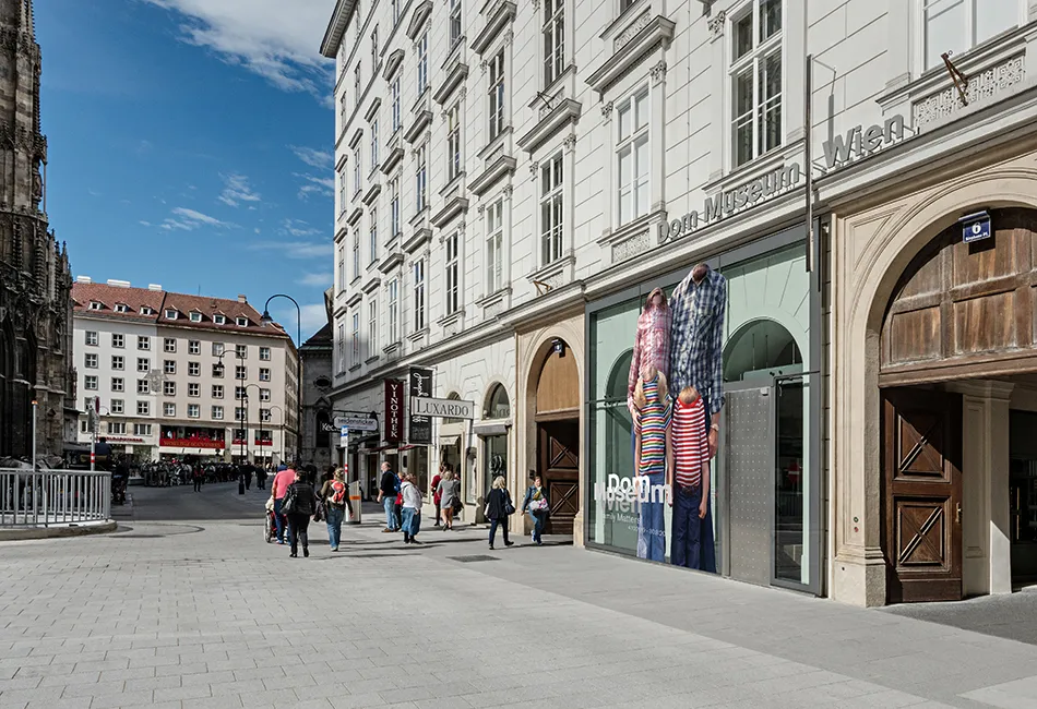 Budynek Dom Museum Wien z szklaną fasadą i dużym oknem wystawowym ukazującym manekiny w tradycyjnych strojach, położony przy ulicy z przechodniami i zabytkowym budynkiem w tle.