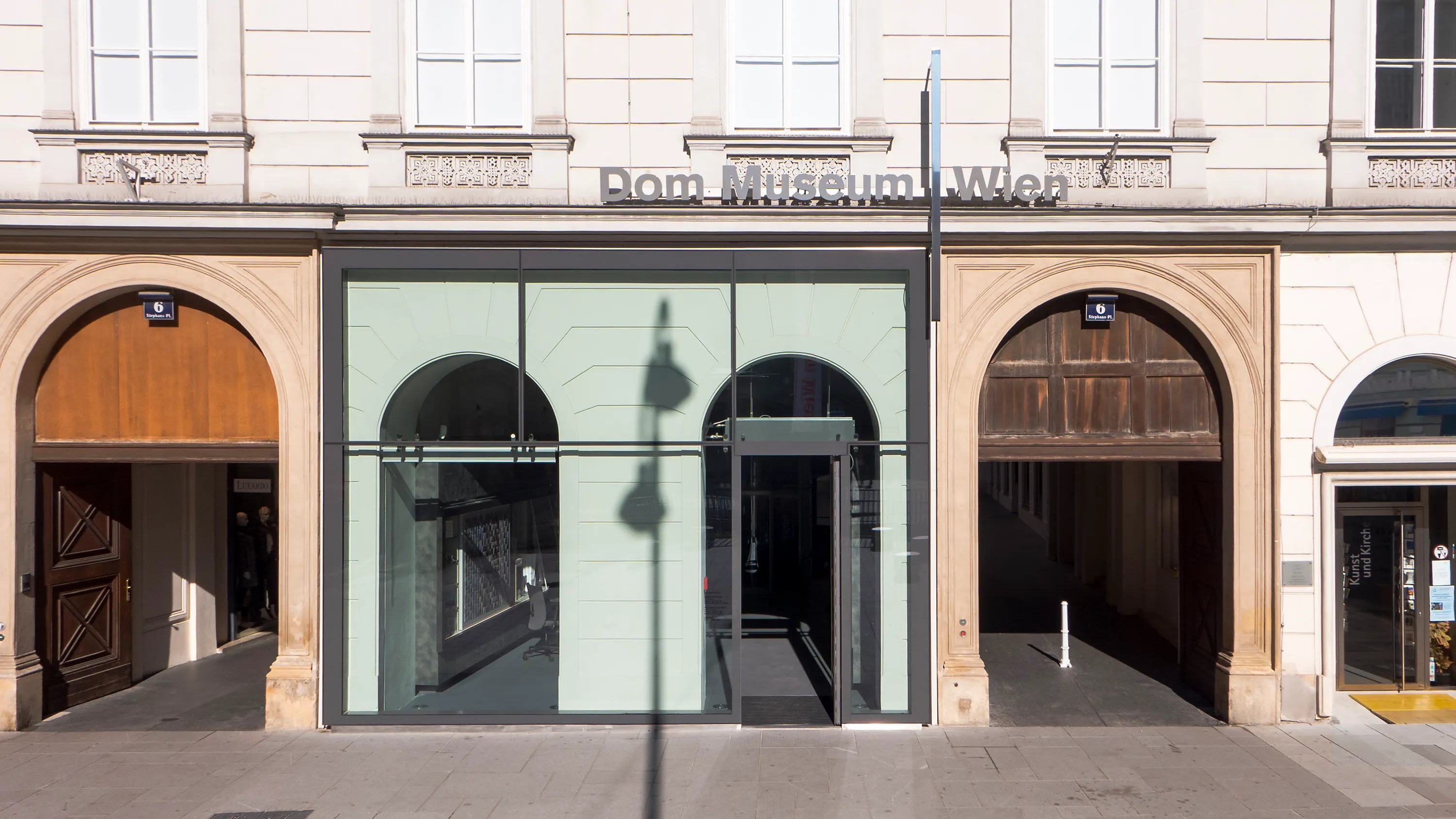 Het gebouw van het Dom Museum Wien met een moderne glazen ingang geflankeerd door gewelfde stenen deuropeningen en een uithangbord boven de ingang.