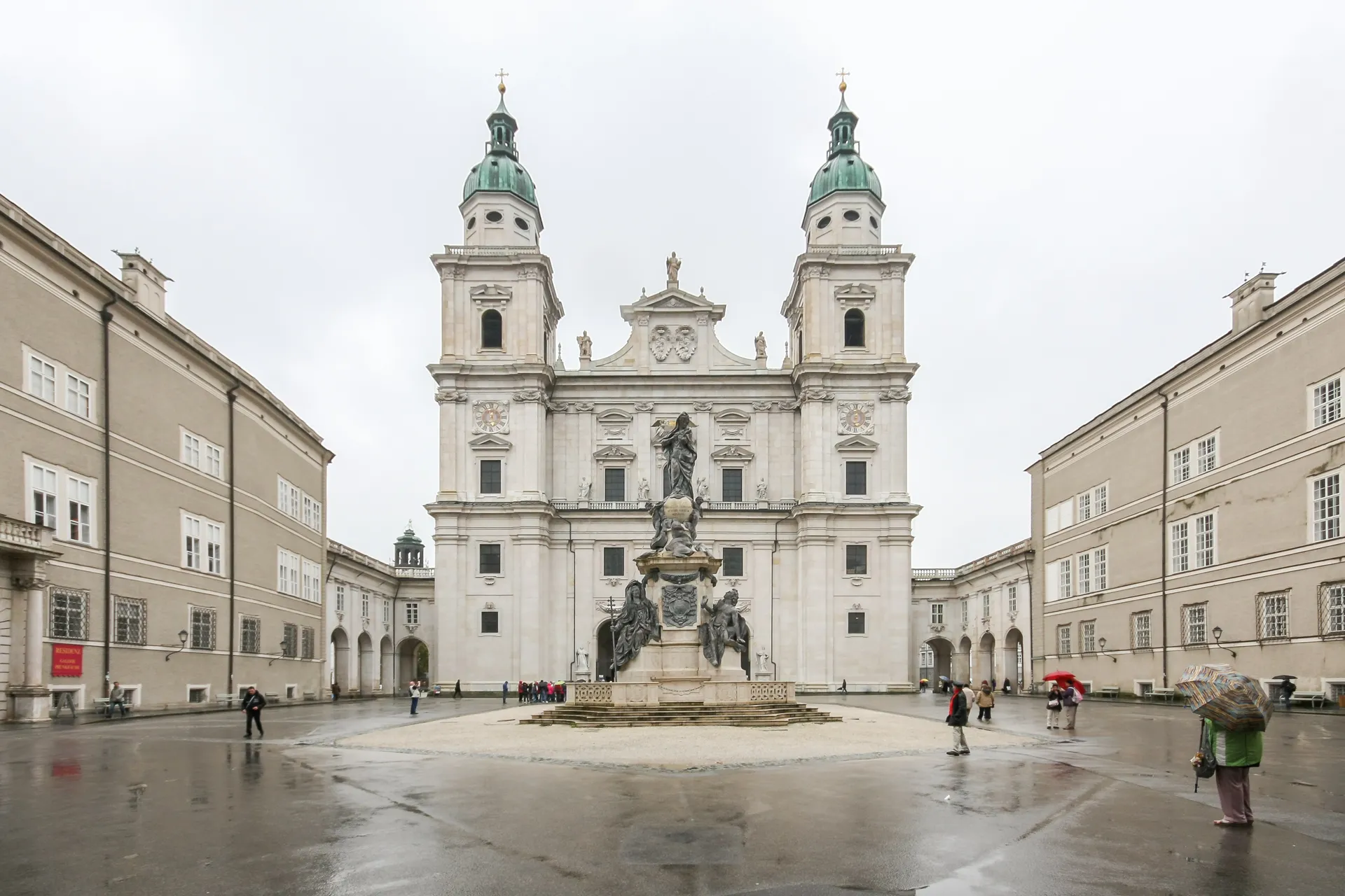 Wide view of Salzburg Cathedral with twin green-domed towers and Marienbrunnen fountain in the center of Residenzplatz square, flanked by arcaded buildings on an overcast day.