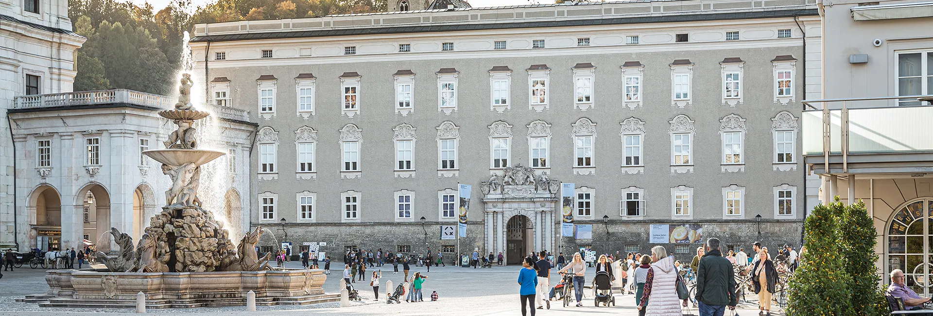 Wide panoramic view of the Salzburg Residenz facade with the ornate Residenzbrunnen fountain in the foreground and visitors walking across the square.