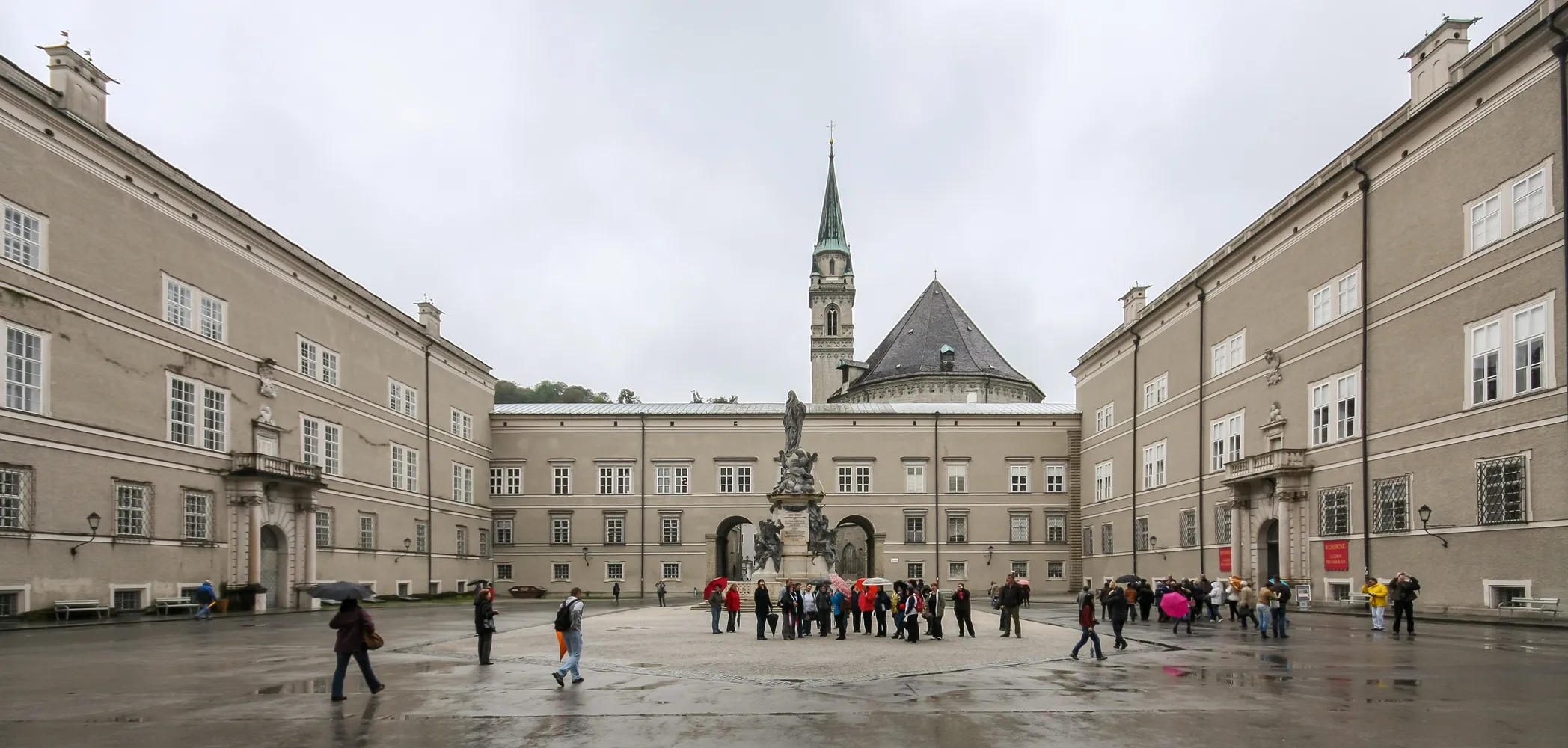 Wide view of the Domplatz courtyard in Salzburg with surrounding baroque buildings, a central fountain with statue, and a church spire in the background, with visitors walking on wet pavement under an overcast sky