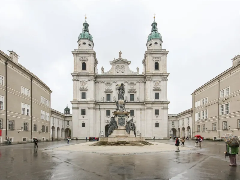 White baroque cathedral facade with two green-domed towers flanking a central section, with a large ornate fountain in the wet plaza in front and people walking around.