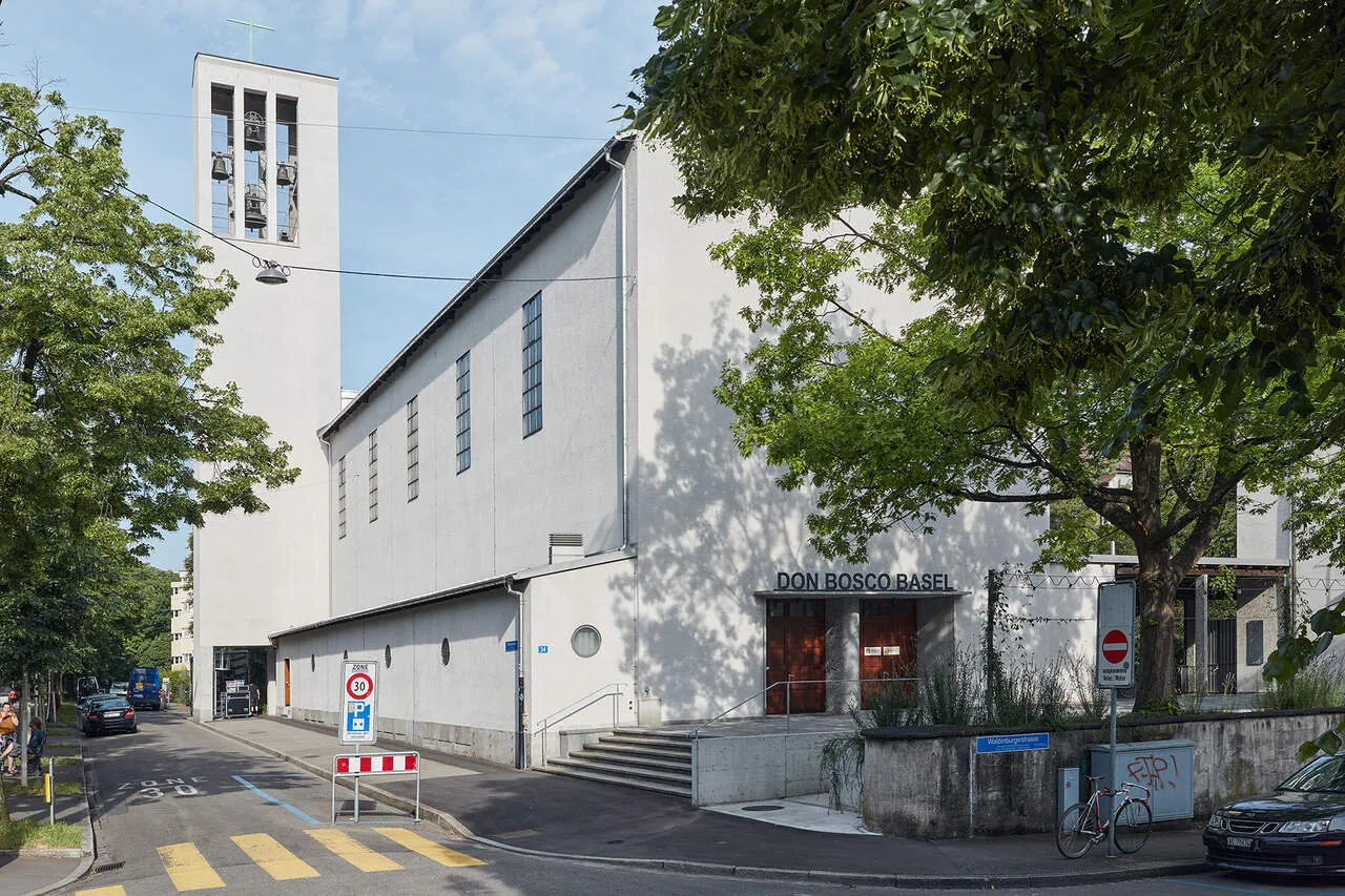 Biały modernistyczny budynek z wysoką dzwonnicą i napisem 'Don Bosco Basel' nad wejściem, otoczony zielonymi drzewami i ulicą z przejściem dla pieszych