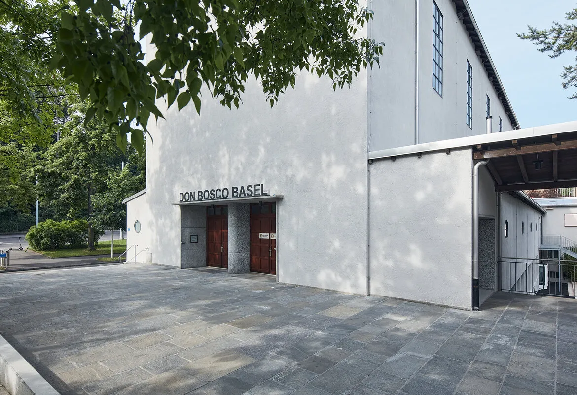 Biała elewacja budynku z napisem DON BOSCO BASEL nad drewnianymi drzwiami wejściowymi, wybrukowany dziedziniec i zielone drzewa na pierwszym planie.