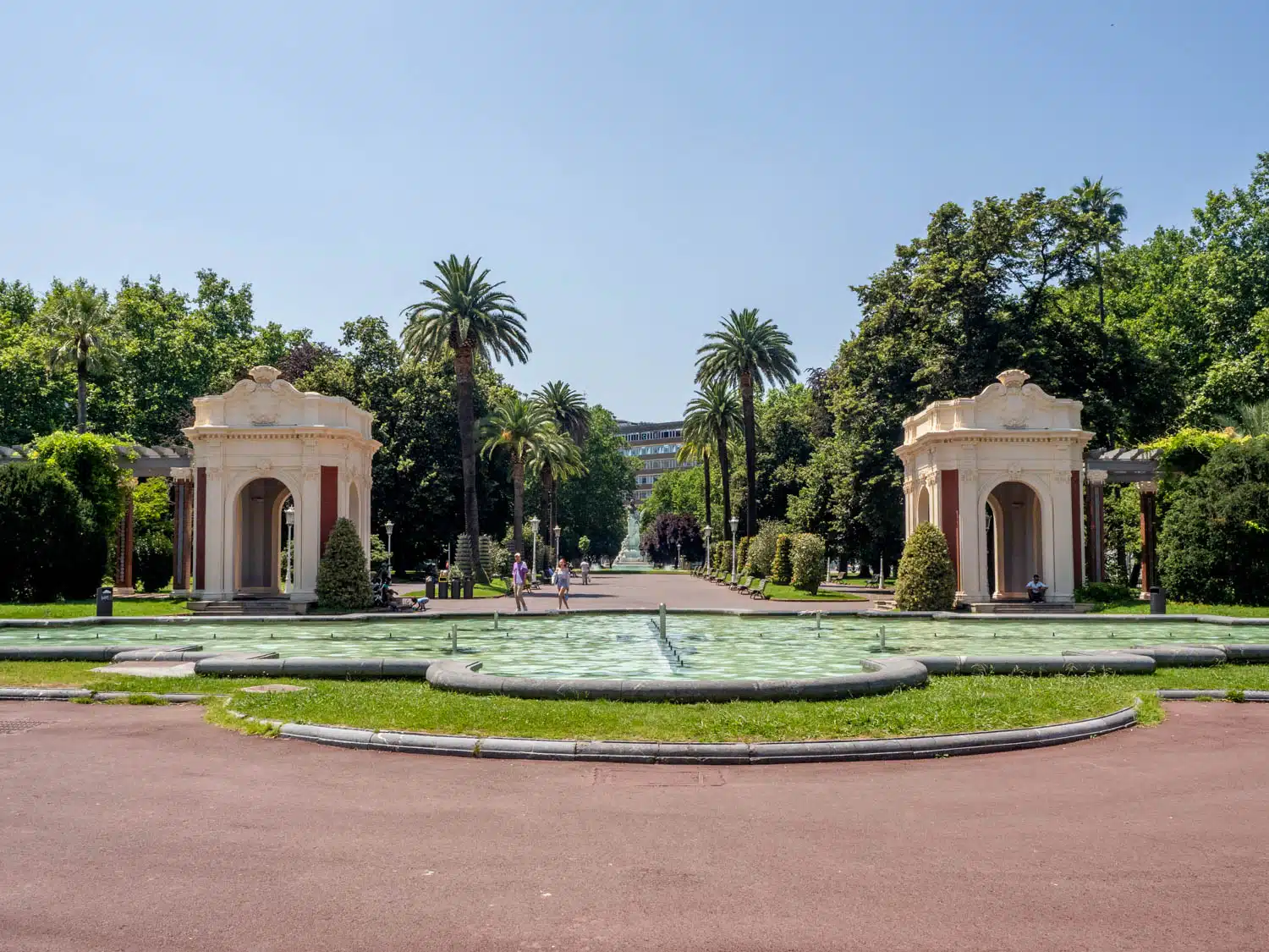 Vista simétrica de la entrada al Parque de Doña Casilda con dos pabellones neoclásicos de color crema flanqueando un camino central, una fuente circular en primer plano y altas palmeras bajo un cielo azul despejado.