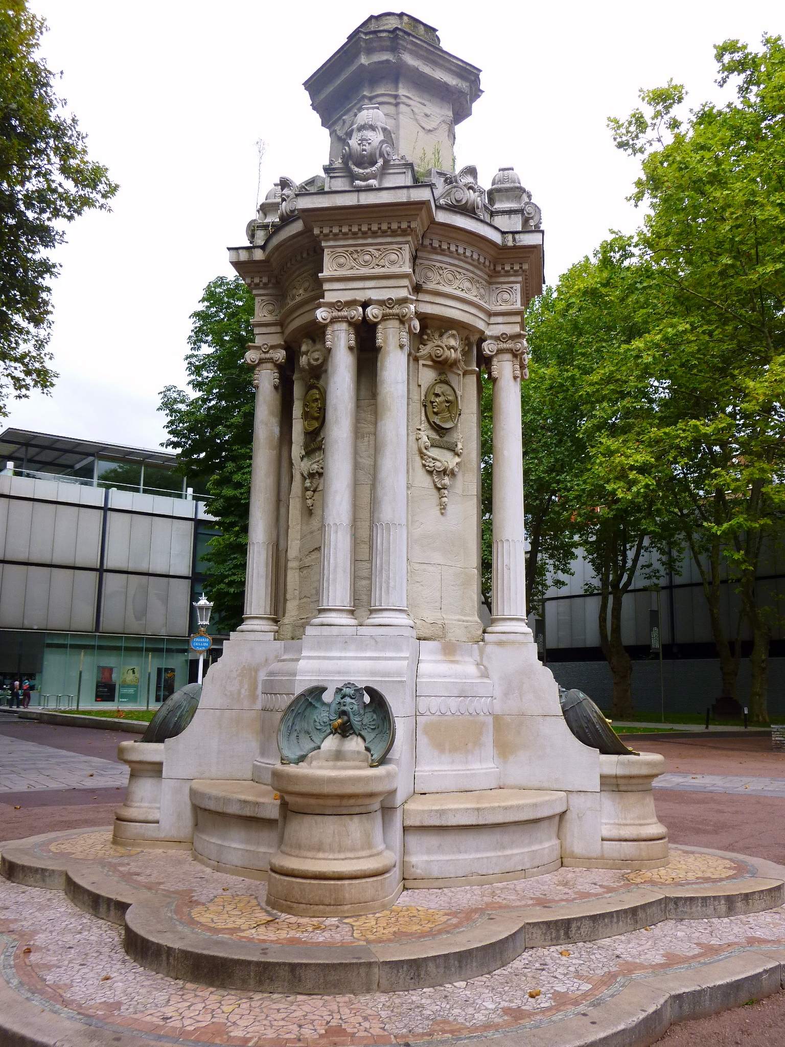 Monumento de fuente de piedra ornamentada con columnas, relieves decorativos y esculturas de águilas de bronce en la base, rodeado de árboles en una plaza del parque