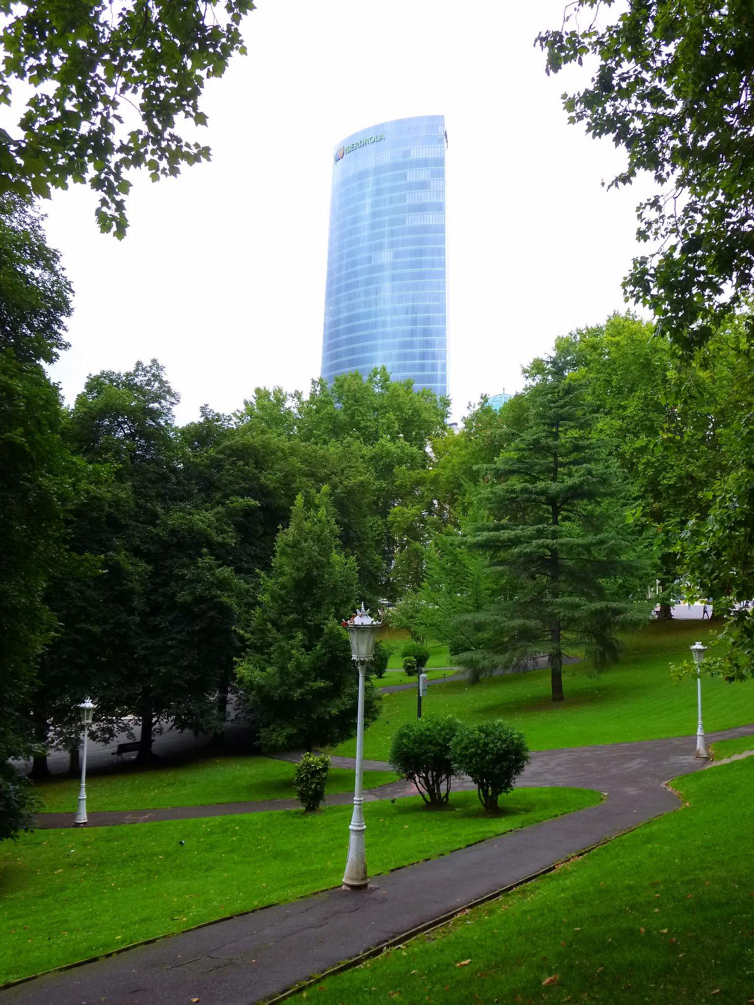 Senderos bordeados de árboles en el parque con farolas blancas y césped verde, con un rascacielos de cristal alto y curvado que se eleva sobre los árboles al fondo