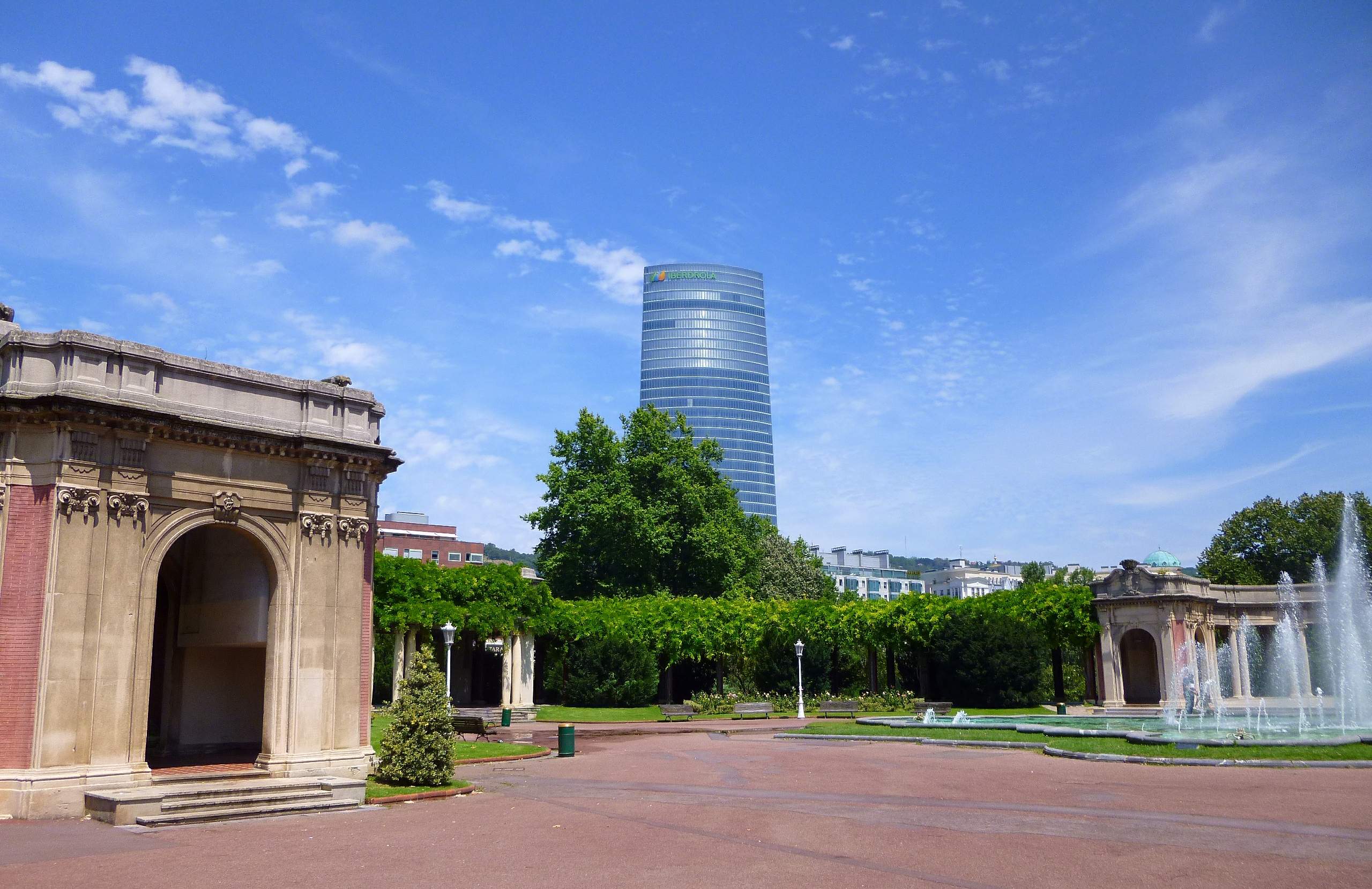 Vista panorámica del Parque de Doña Casilda, que muestra estructuras clásicas de piedra, una fuente, setos bien cuidados y un rascacielos de cristal moderno al fondo bajo un cielo azul.
