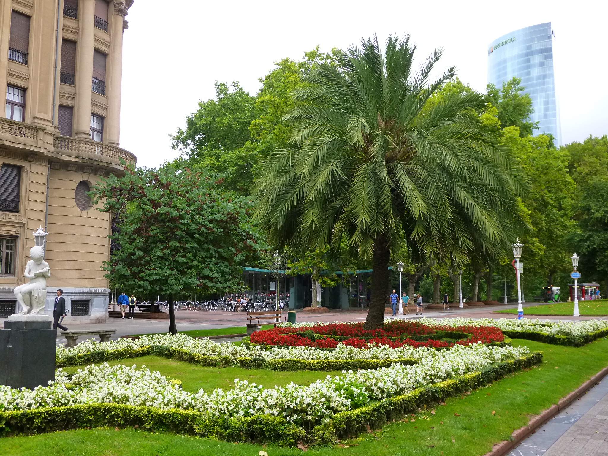 Jardín paisajístico con una gran palmera central rodeada de parterres de flores blancas y rojas y setos cuidados, flanqueado por un edificio clásico de piedra y un moderno rascacielos de cristal al fondo.