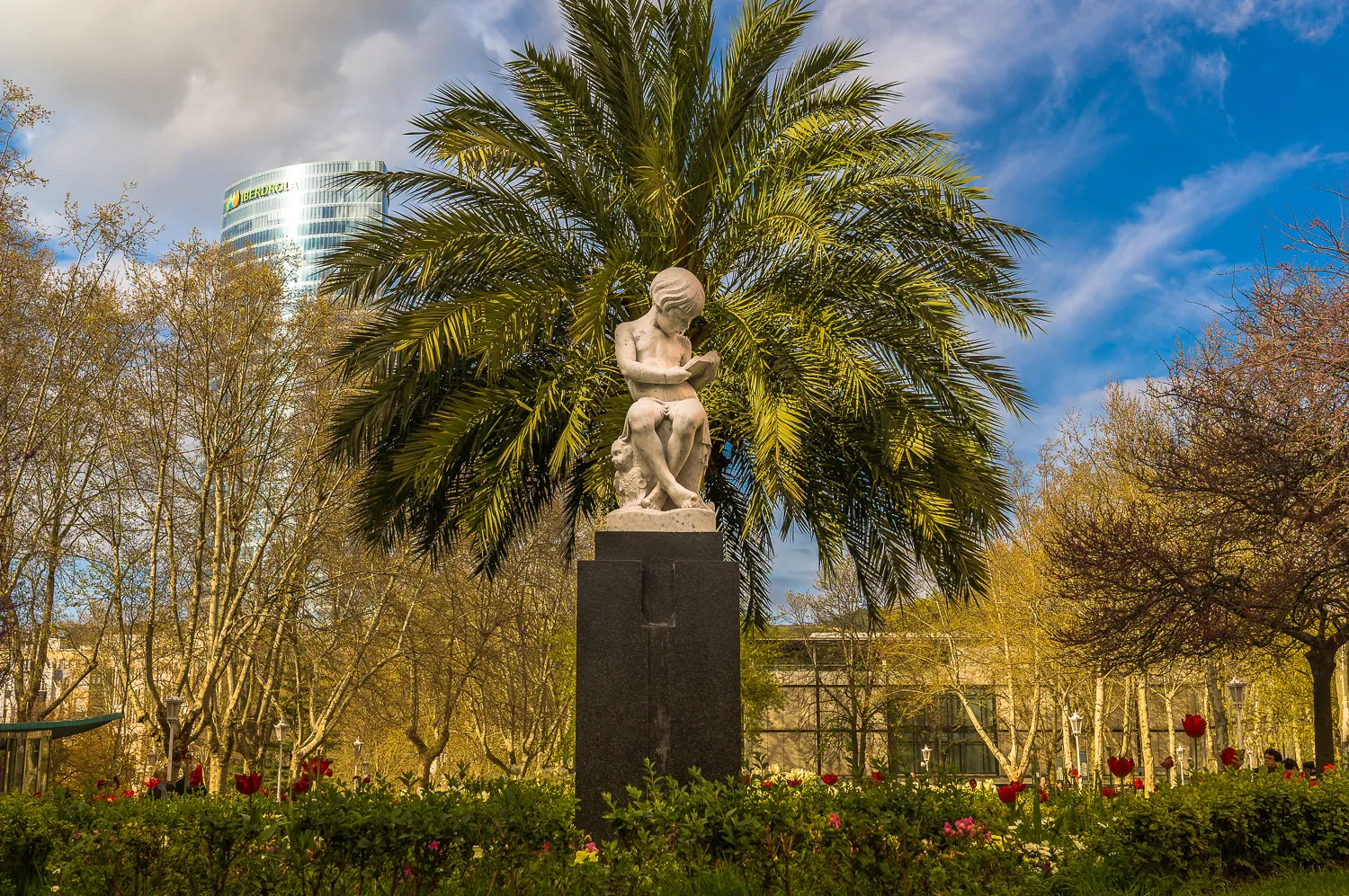 Una estatua de mármol blanco de una figura sentada sobre un pedestal oscuro, enmarcada por una gran palmera, con parterres de flores rojas y un moderno edificio de cristal al fondo.