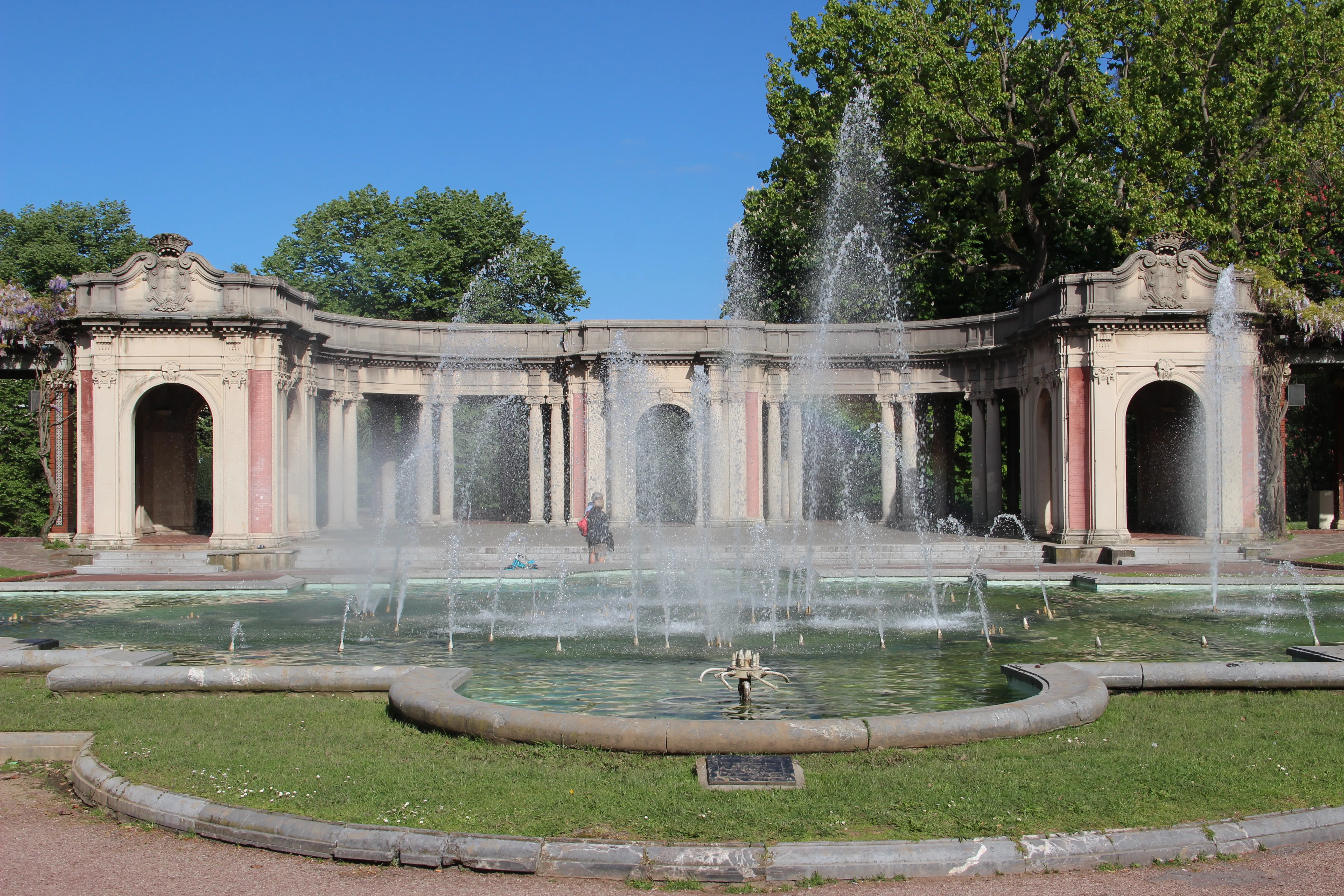Una toma panorámica de una columnata de piedra curva con arcos detrás de una gran fuente con múltiples chorros de agua en un parque en un día soleado.