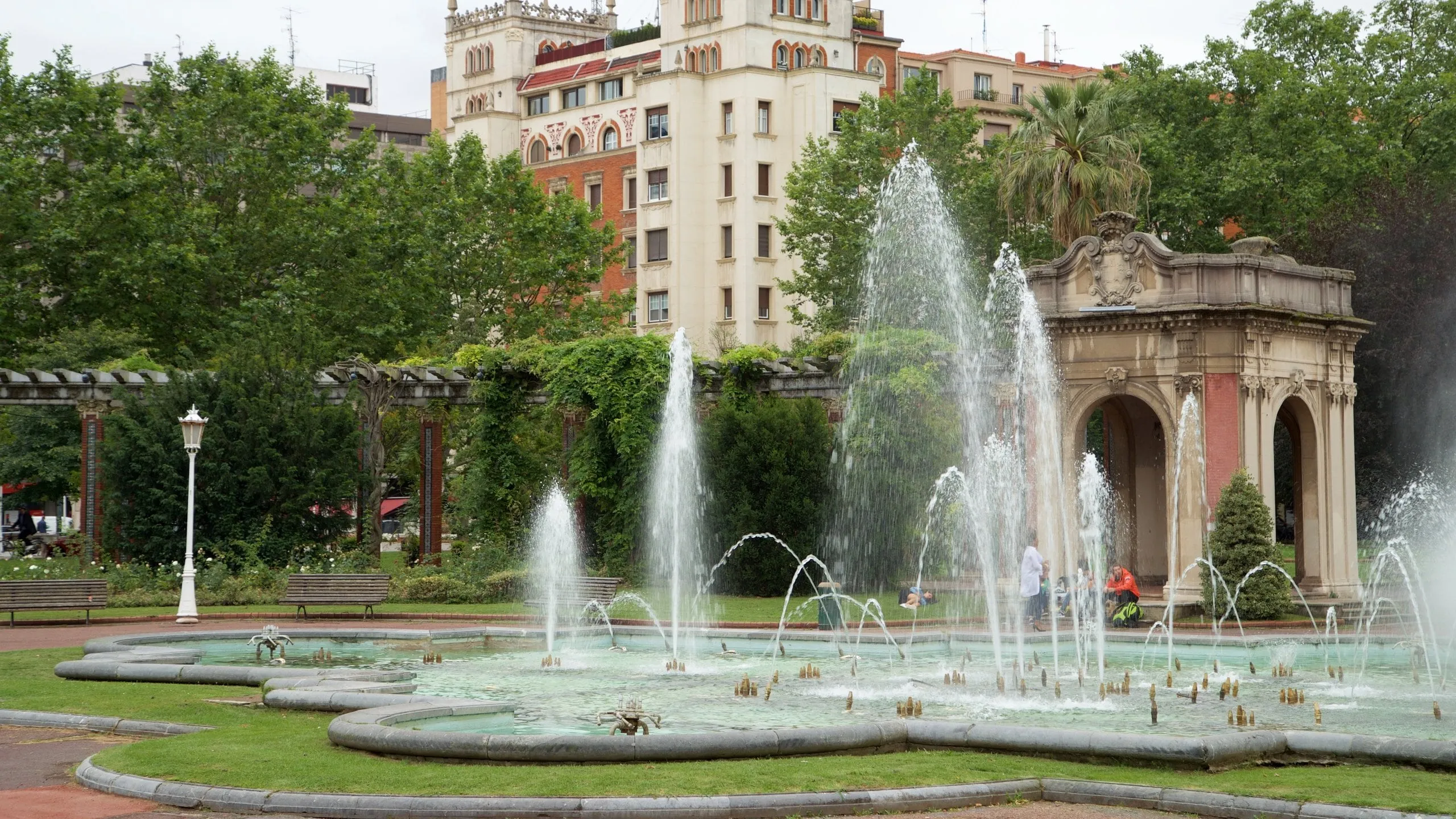 Fuentes de agua en el parque de Doña Casilda con un pabellón de piedra clásico con arcos, una pérgola de madera, zonas verdes, árboles y edificios de apartamentos al fondo.