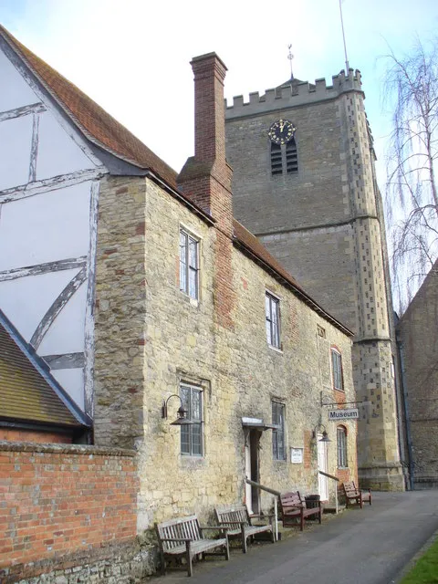 Stone building with a Museum sign next to a church tower with a clock, wooden benches along the path