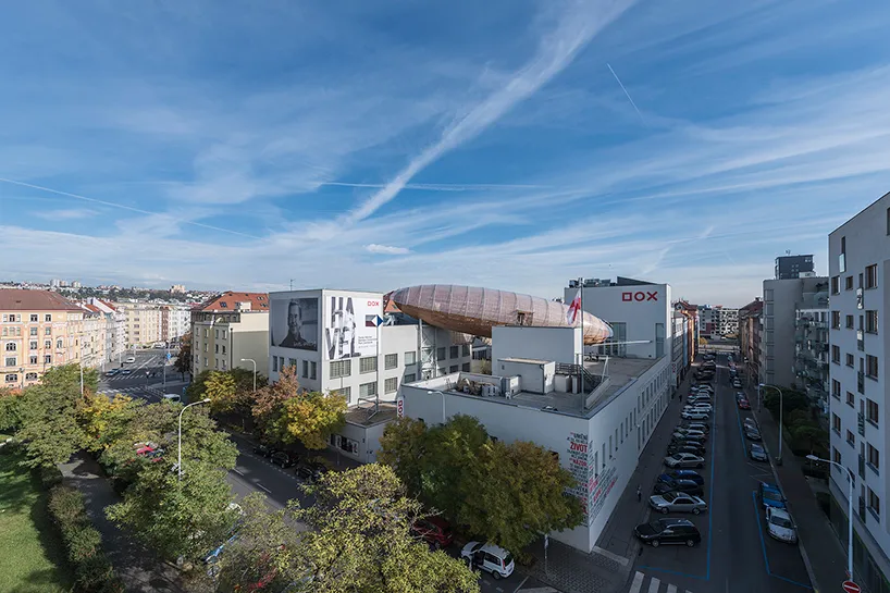 Widok z lotu ptaka na Centrum Sztuki Współczesnej DOX z dużą rzeźbą sterowca na dachu, otoczone miejskimi budynkami i drzewami pod błękitnym niebem.