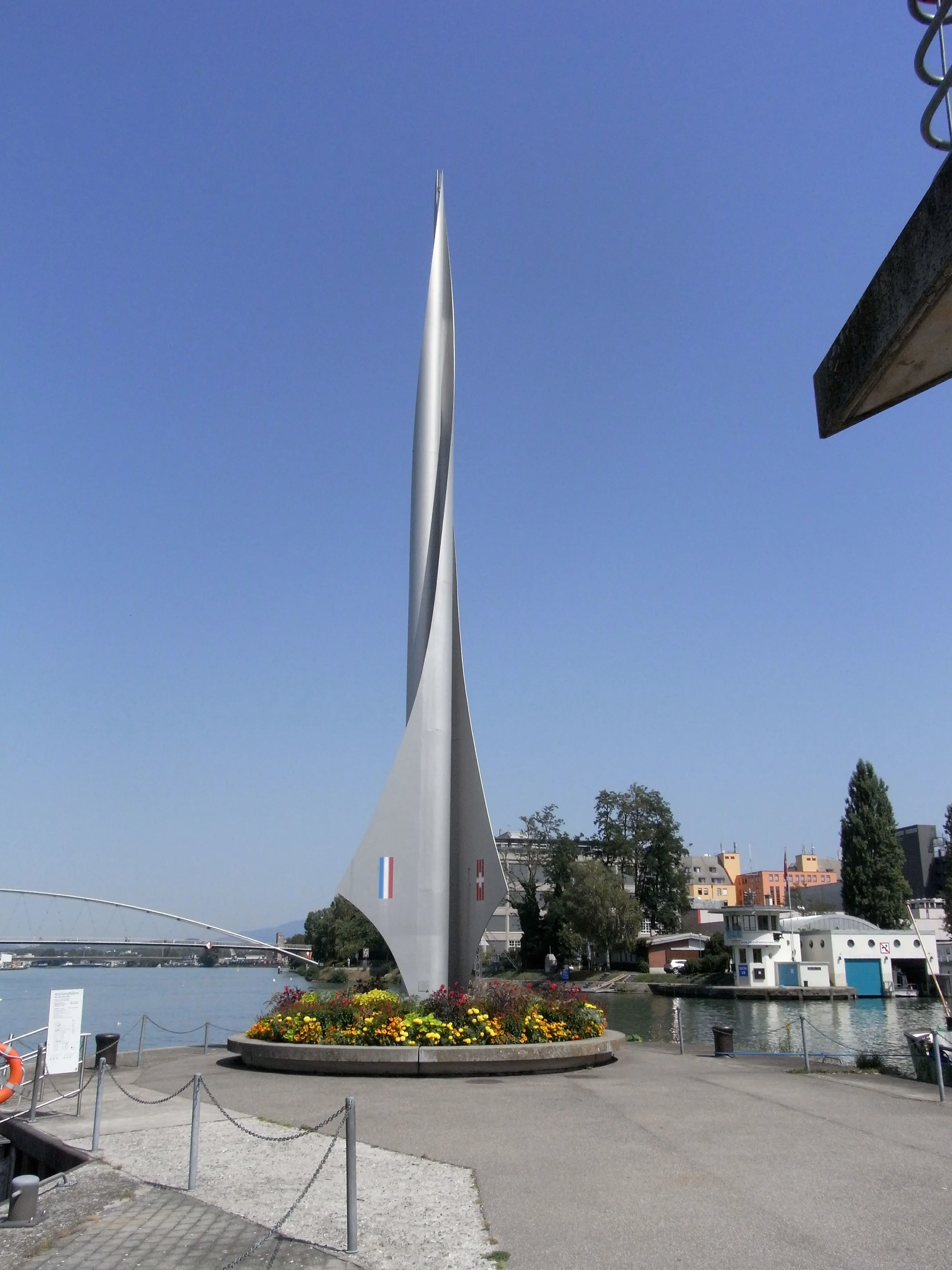Tall silver rocket-shaped Dreiländereck monument rising from a circular flower bed near the Rhine river, with French, German, and Swiss flags at its base and harbour buildings in the background