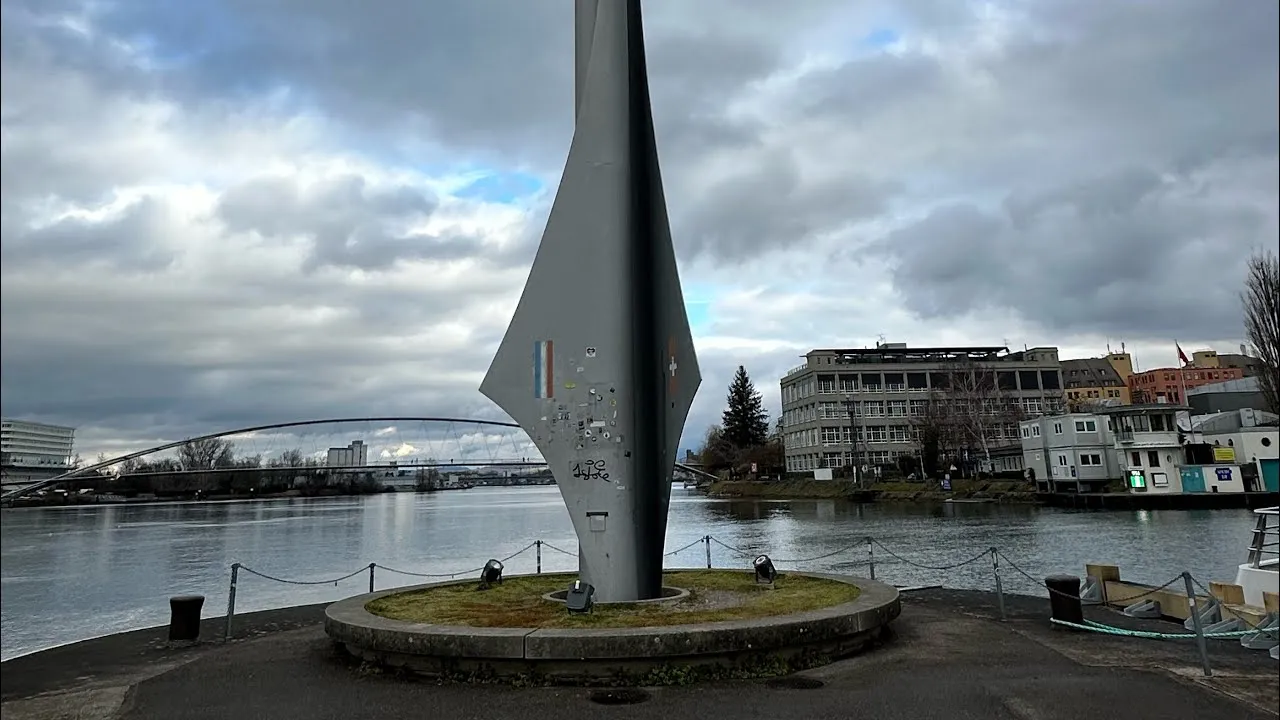 Tall grey rocket-shaped monument on a circular base near the Rhine river with a bridge and buildings in the background under a cloudy sky.