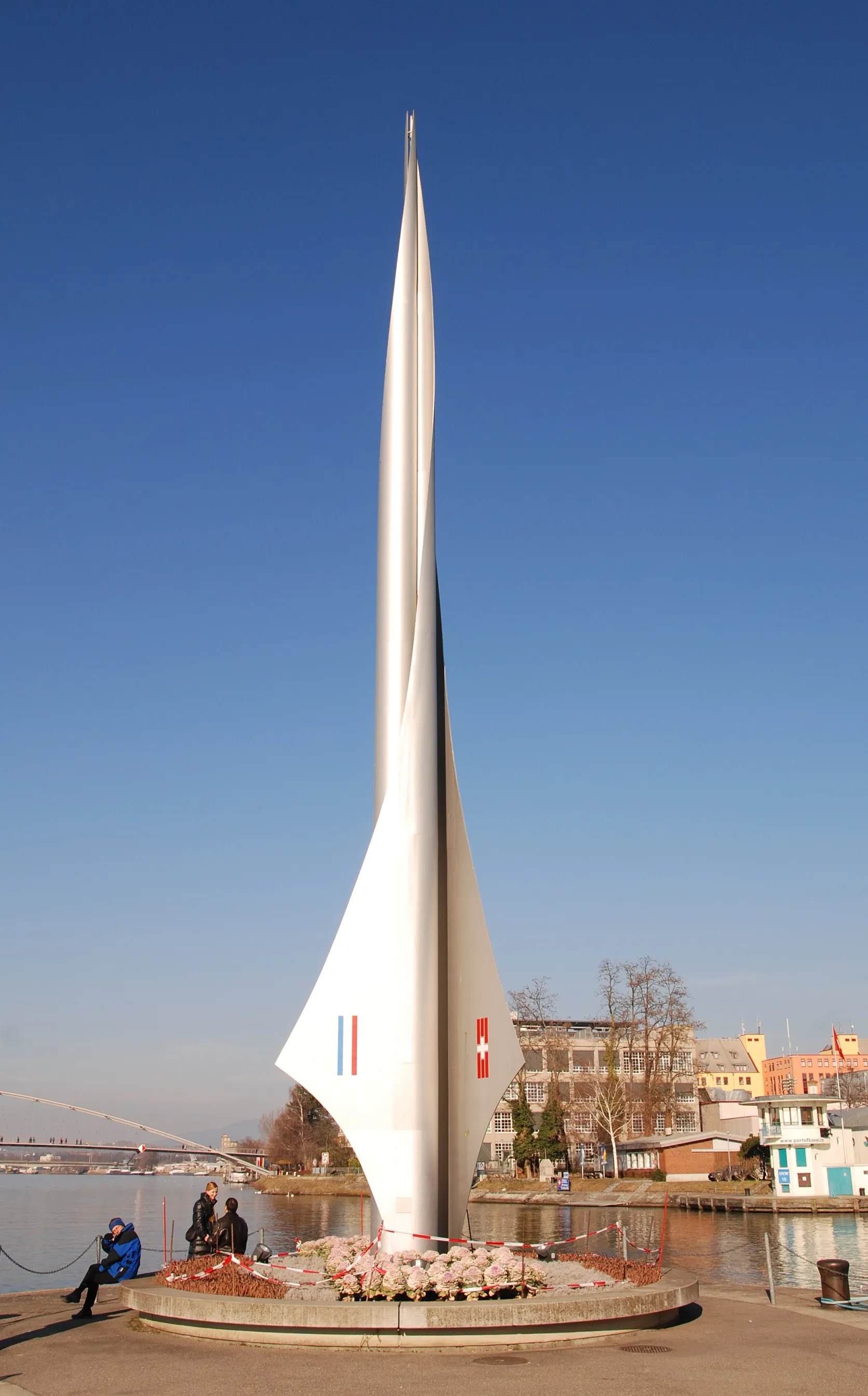 A tall, white, sail-shaped monument displaying the flags of France, Switzerland, and Germany, standing on a circular base near a river with people sitting nearby under a clear blue sky.