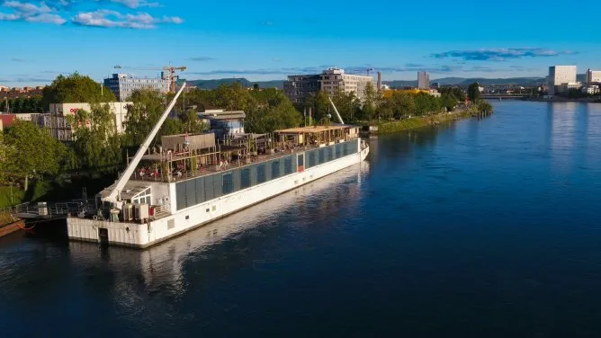 Aerial view of a long white river barge docked along the Rhine riverbank with a crane, tree-lined shore, and city buildings under a blue sky