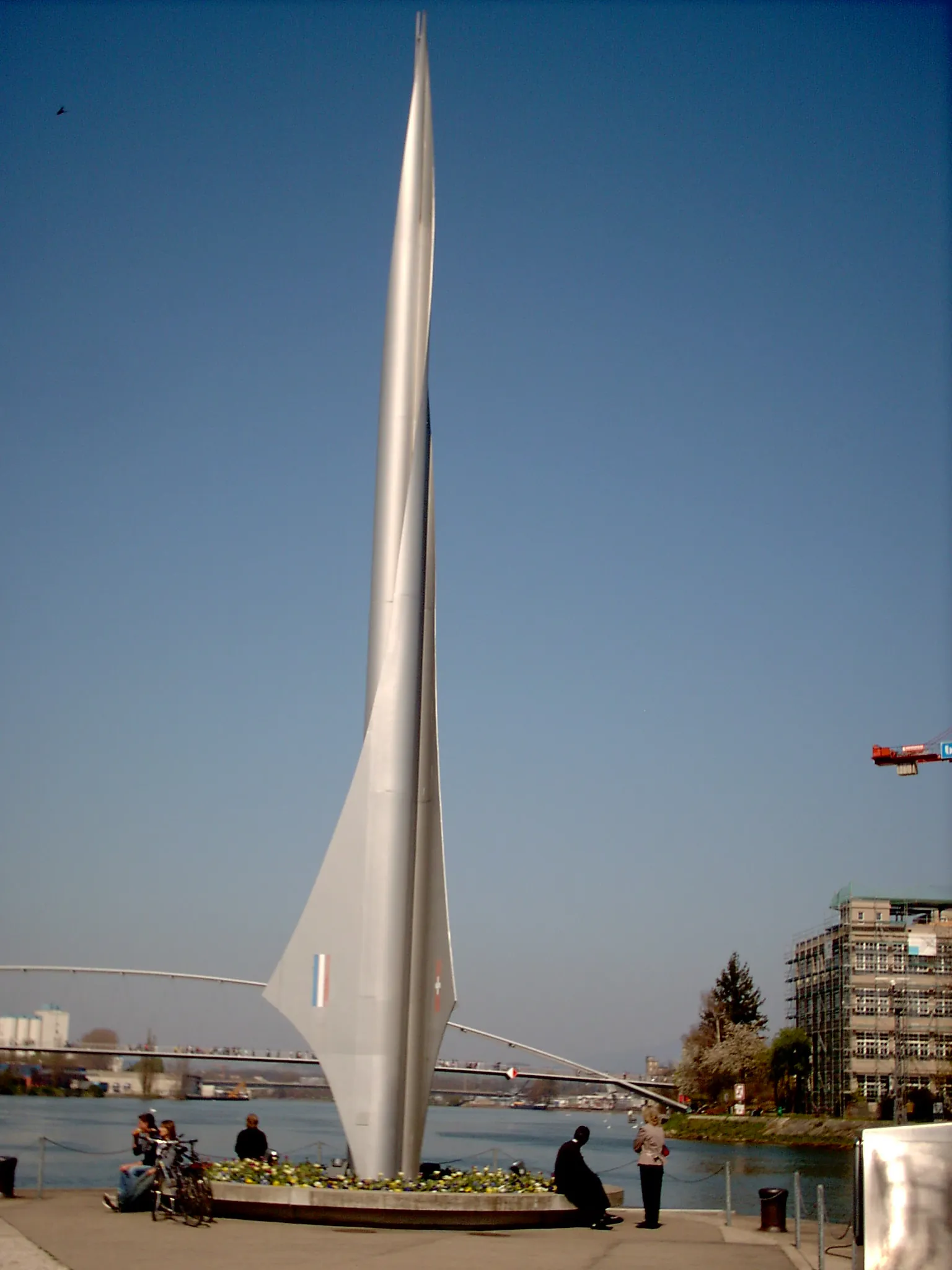 Tall silver sail-shaped monument rising from a circular flower bed at the riverbank, with people sitting and standing nearby, the Rhine river and a bridge visible in the background under a clear blue sky.