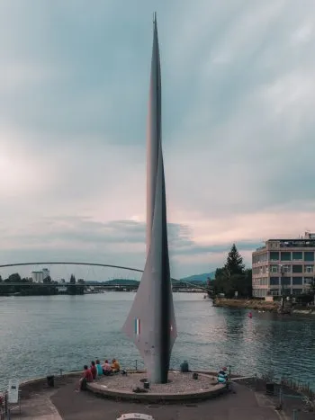 A tall, slender, rocket-shaped monument rises from a circular platform on a river pier, with a few people sitting at its base and an arched bridge spanning the water in the distance.