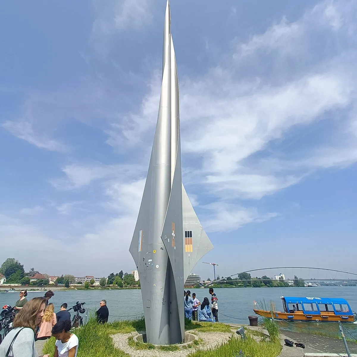 Tall silver rocket-shaped Dreiländereck Monument standing on a grassy peninsula beside the Rhine river, with visitors gathered around its base and a blue tour boat on the water
