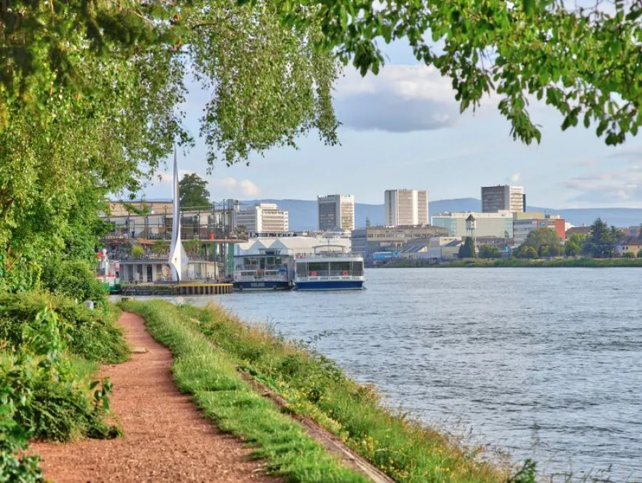 Gravel path along the Rhine river with the white Dreiländereck Monument spire near a dock, moored boats, green trees overhead, and Basel city buildings across the water