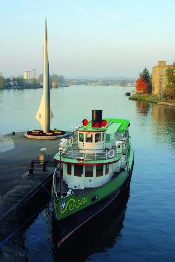 Rocket-shaped white Dreiländereck monument standing on a dock beside a green and white paddle steamer boat on the Rhine river in Basel