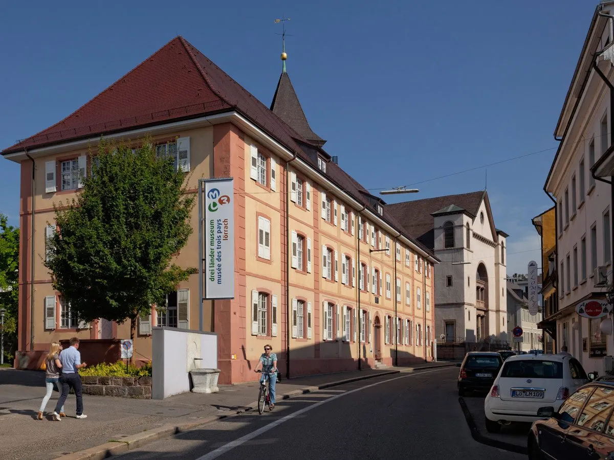 Buitenaanzicht van het Dreiländermuseum, een groot rozeachtig historisch gebouw met een donkerrood dak en een verticaal spandoek met de museumnaam, terwijl mensen op straat wandelen en fietsen.