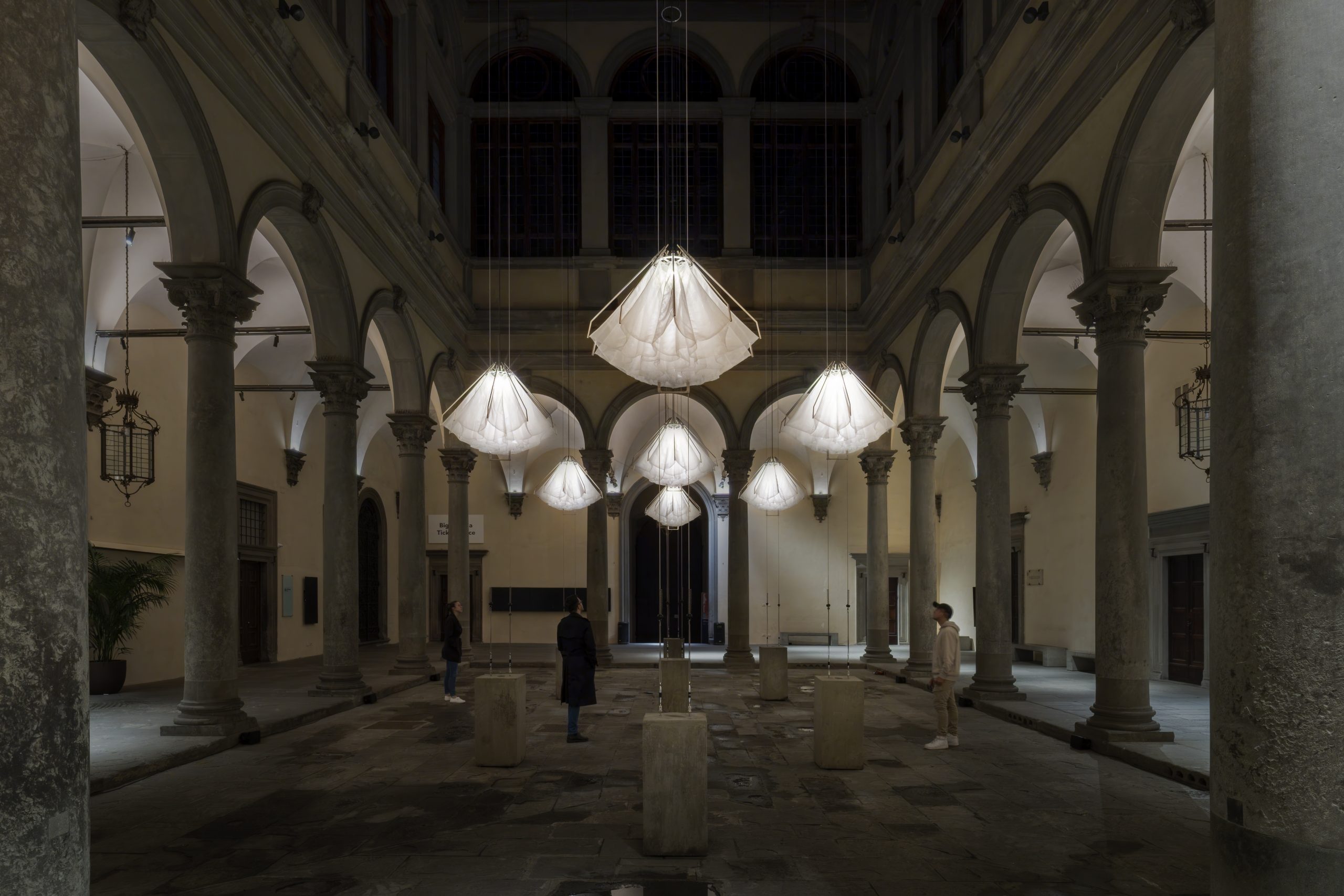 Dimly lit grand hall with stone arches and suspended illuminated light sculptures; visitors walk among cylindrical platforms