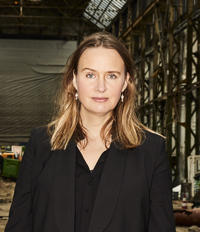 A person in a black suit stands in an industrial interior space with metal beams and a high ceiling