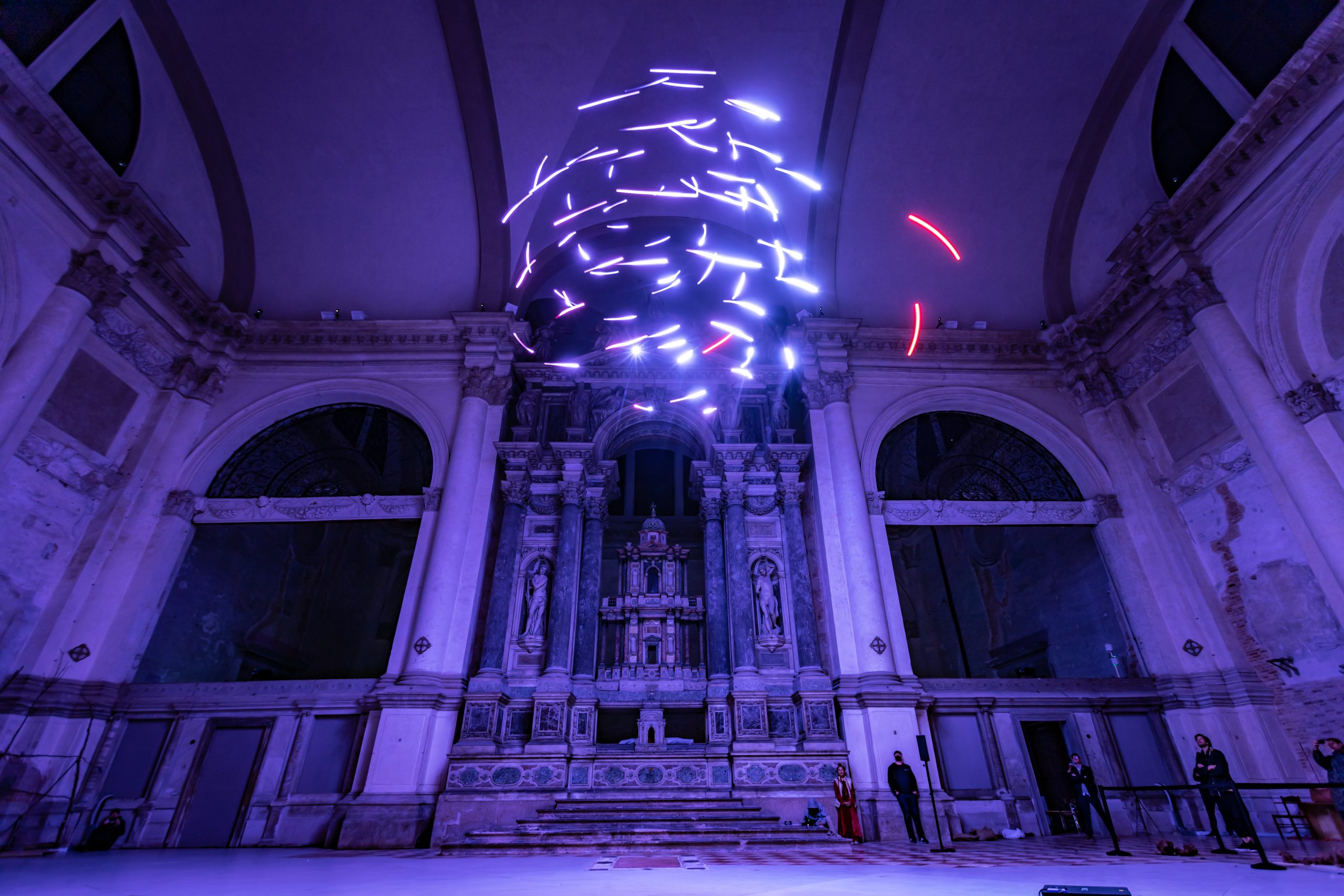 Immersive light installation with glowing elements suspended in a grand, arched hall with statues and visitors observing