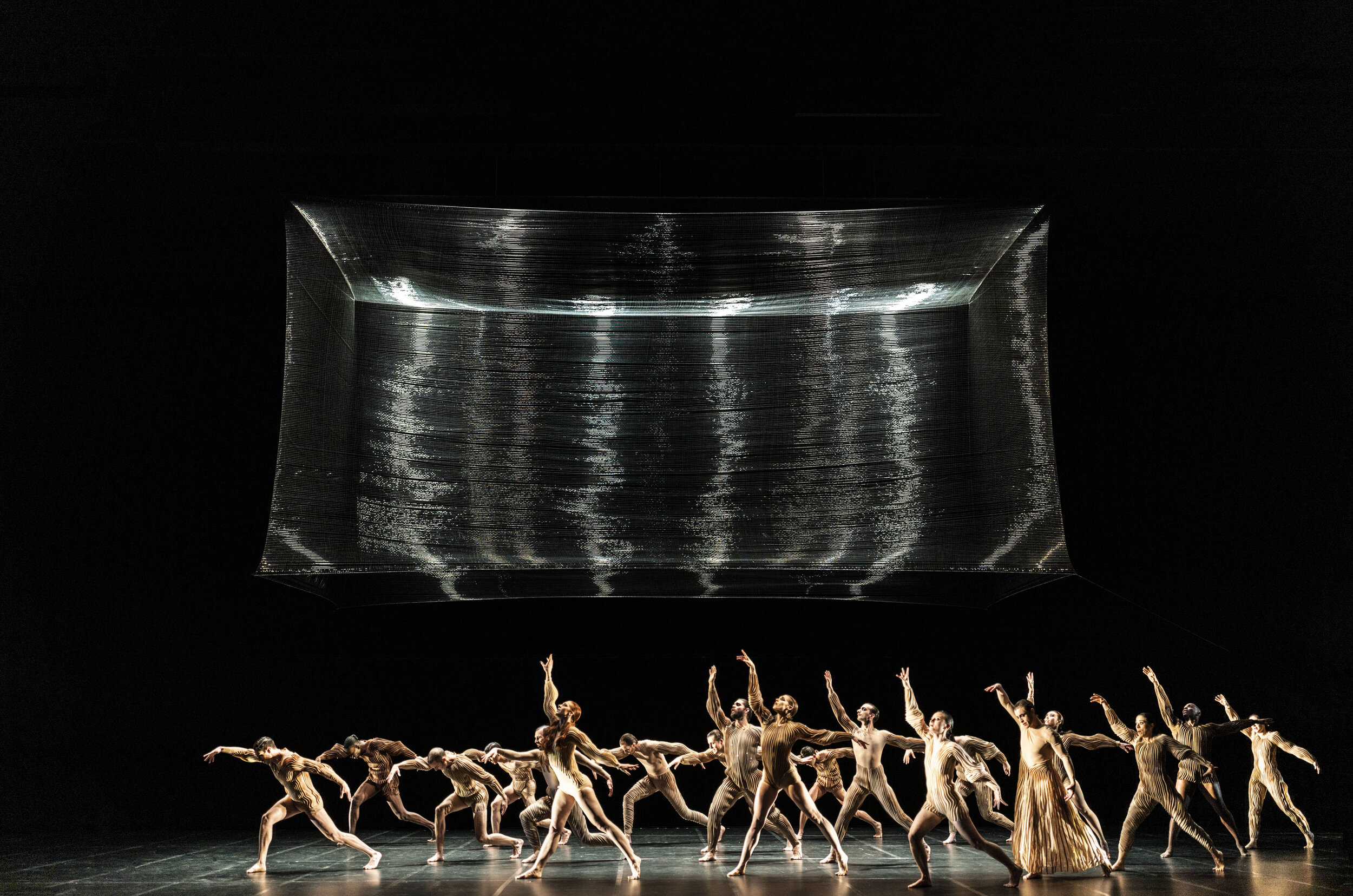Dancers perform on stage beneath a large digital display showing rippling water patterns in a dark theater setting