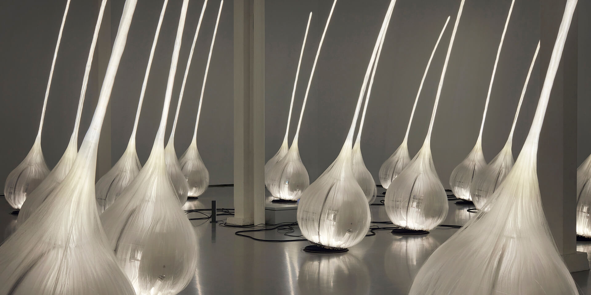 White elongated bulbous light sculptures arranged in rows within a dimly lit gallery space, part of an immersive digital art installation.