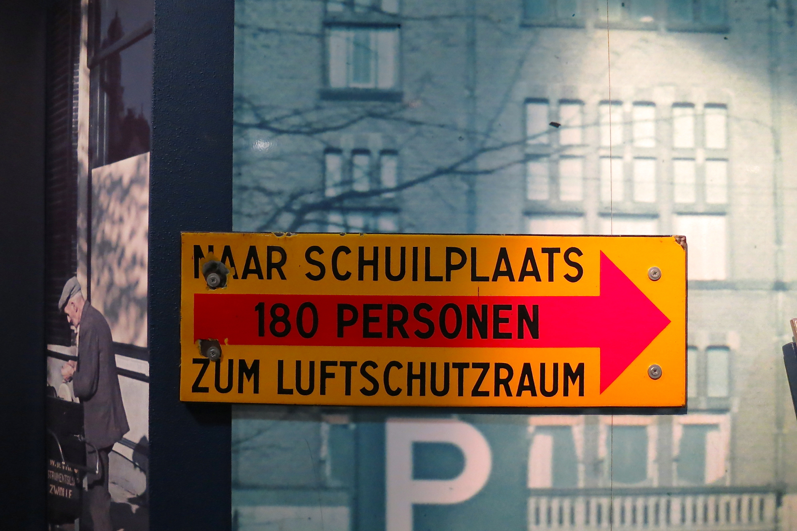 Geel en rood richtingbord met Nederlandse en Duitse tekst 'NAAR SCHUILPLAATS 180 PERSONEN ZUM LUFTSCHUTZRAUM' en pijl, tentoongesteld in het Verzetsmuseum
