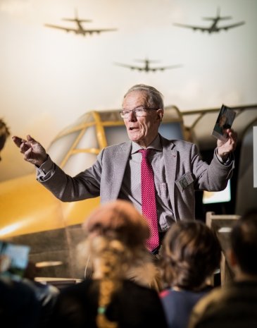 A man in a gray suit and red tie speaks to an audience in front of a yellow aircraft display with military plane silhouettes