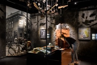 Donker verlichte museumexpositie met historische foto's, een fiets en een persoon die artefacten onderhangende houten balken onderzoekt
