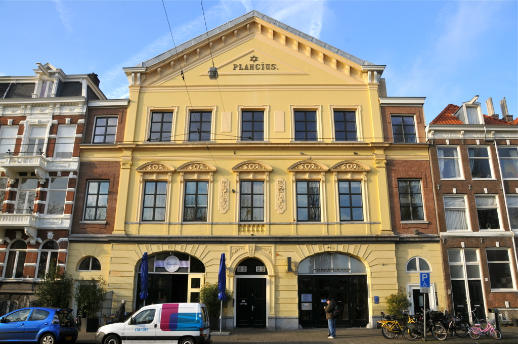 Geel historisch gebouw met 'PLANCius'-bord, rondboogvensters en ingang met fietsen en voertuigen die buiten op een zonnige dag geparkeerd staan