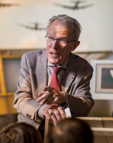 Man in gray suit with red polka-dot tie speaking in museum room with airplane silhouettes on wall and audience members visible