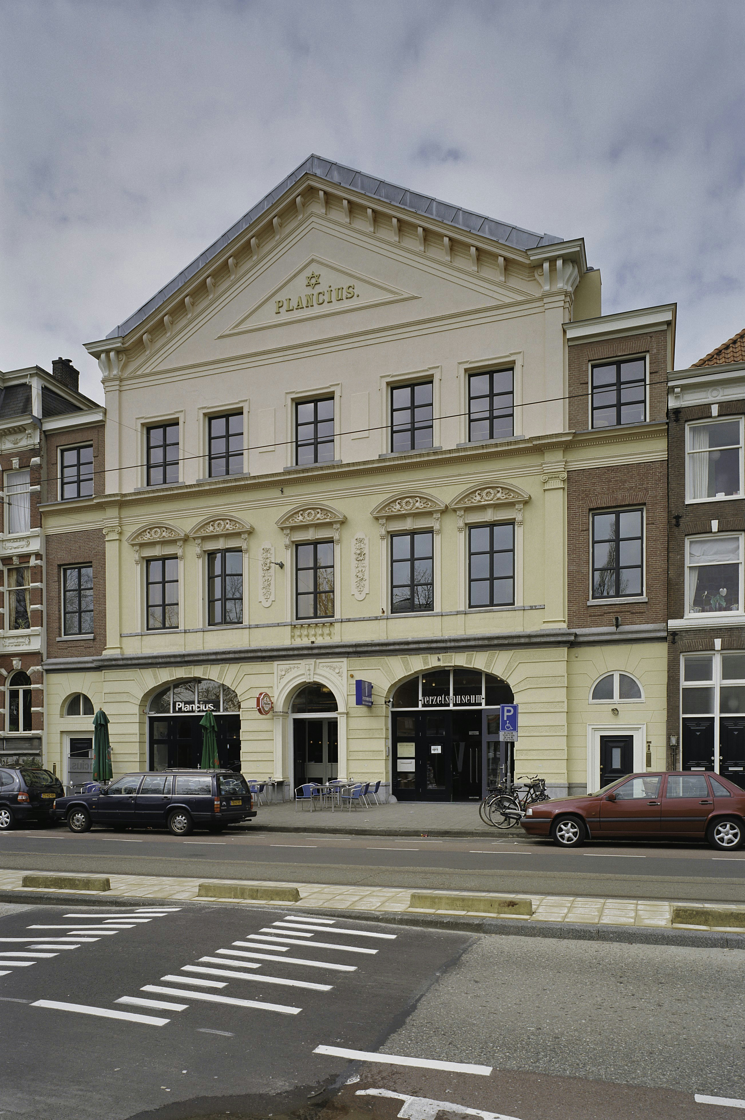 Een bleekgeel gebouw met een driehoekig fronton en bovenaan het woord 'PLANCUS', met boogvormige ingangen en ramen, geparkeerde auto's ervoor en een zebrapad op de voorgrond.