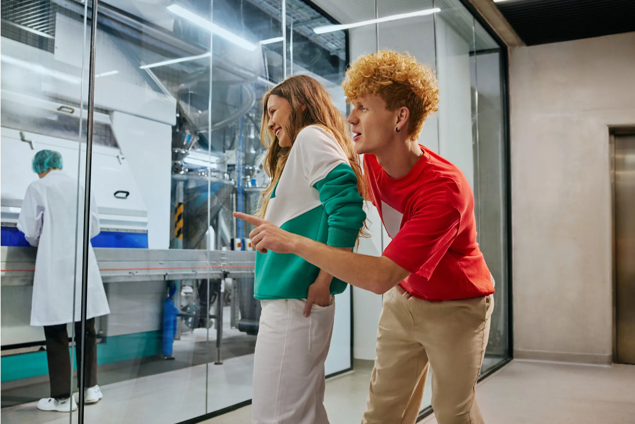 Two visitors looking through a glass wall into a factory production area where a worker in a white coat is operating machinery