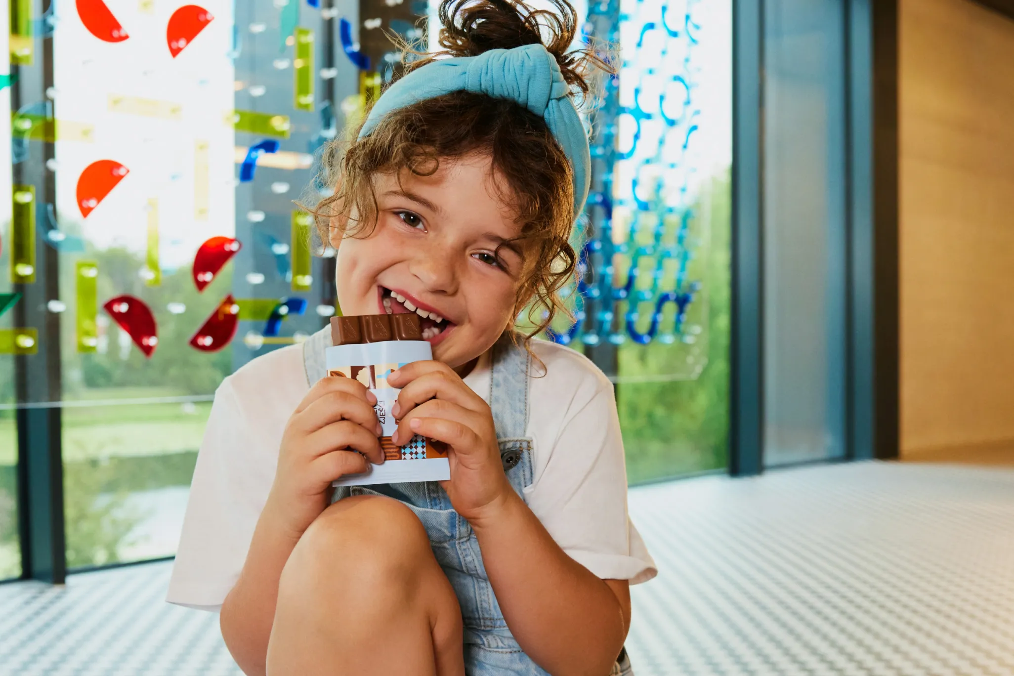 A young girl in overalls and a blue headband holding a chocolate bar and smiling inside a modern space with colorful glass art.