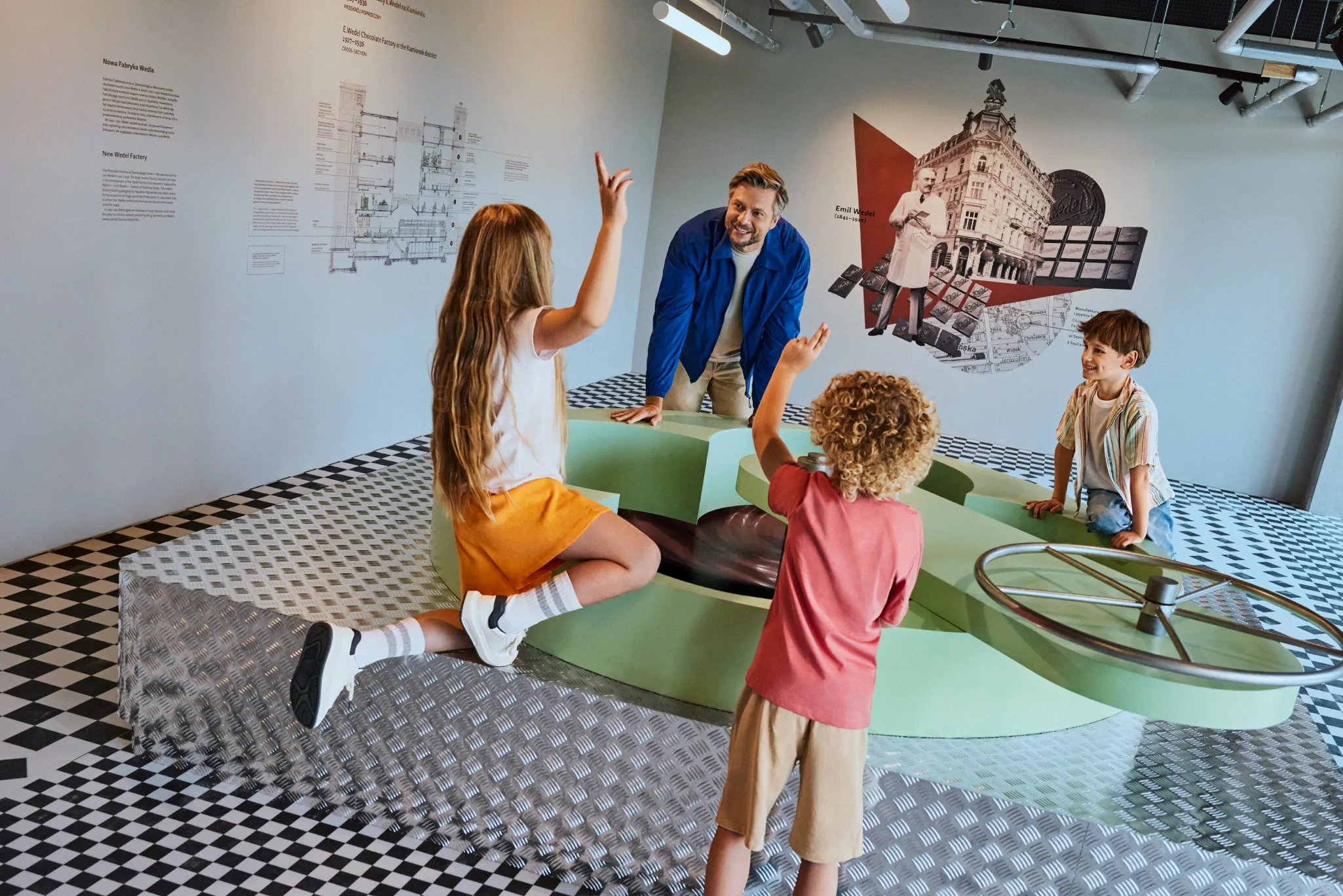 Children and an adult interacting with a green exhibit in a museum with a checkered floor and historical illustrations on the wall