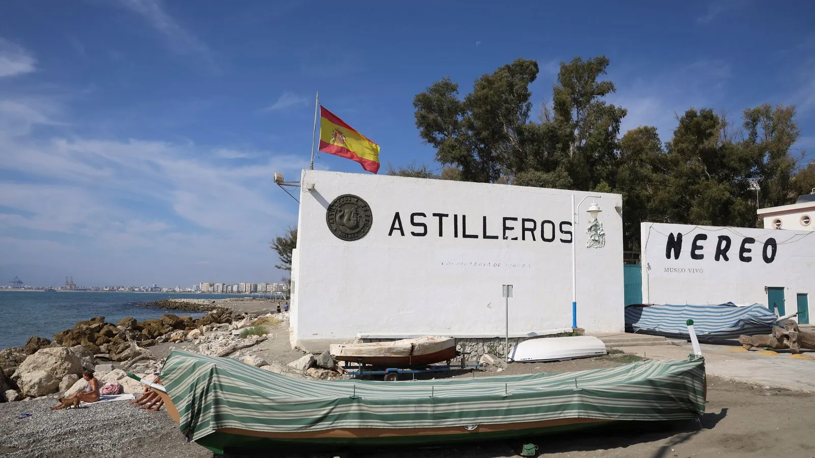 Vista exterior del astillero Astilleros Nereo que muestra paredes blancas con el nombre del lugar en letras grandes, una bandera española ondeando encima y un barco de madera tradicional cubierto con una lona de rayas verdes, con el mar y el horizonte de la ciudad de Málaga de fondo.