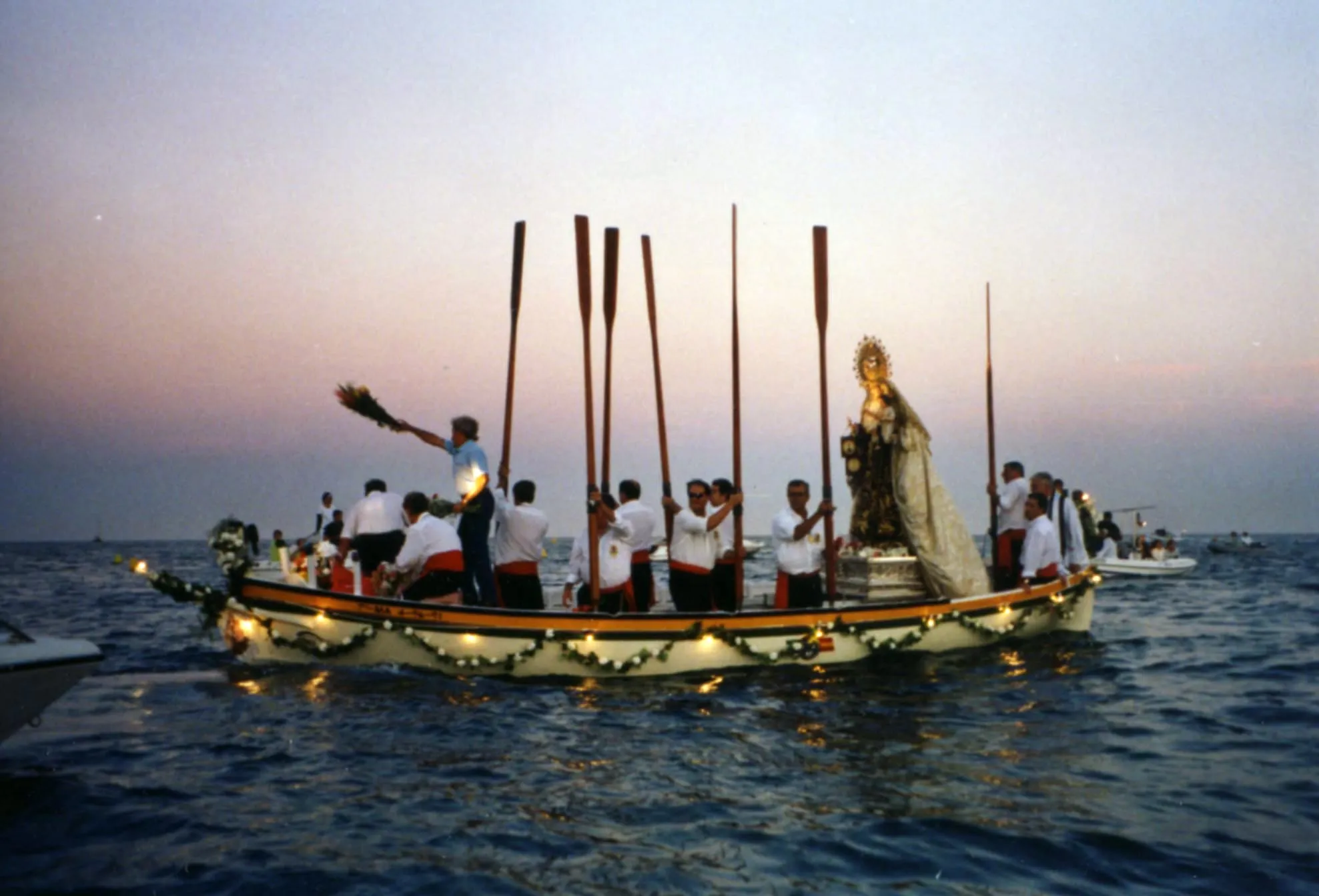 Barco de madera tradicional decorado con guirnaldas de luces, que transporta una estatua religiosa y una tripulación de remeros con remos largos en el mar al atardecer